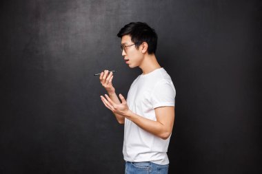 Teknoloji, aletler ve insan konsepti. Asyalı genç erkek öğrencinin profil portresi cep telefonundaki sesli mesaj kaydı, akıllı telefonu dudaklarının yakınında tut, siyah arkaplan