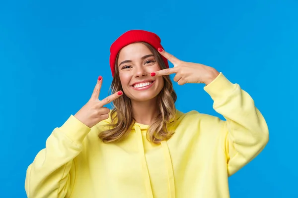 Despreocupada mujer europea guapa en gorro rojo y sudadera con capucha amarilla, sonriente kawaii mostrar gestos de paz como el envío de vibraciones positivas, de pie optimista sobre fondo azul —  Fotos de Stock