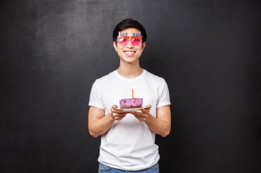 Doğum günü, kutlama ve parti konsepti. Beyaz tişörtlü, komik gözlüklü, elinde mumlu doğum günü pastası olan mutlu, genç, neşeli Asyalı adam. Dilek tutmak için üfle, heyecanlı gülümse, siyah arka planda dur.