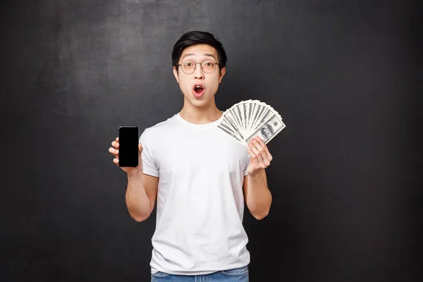 Technology, money and prizes concept. Amazed and surprised asian guy holding dollars, big cash and mobile phone, showing screen of smartphone banking application, look shocked camera