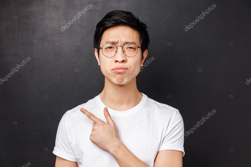 Close-up portrait of picky, skeptical and gloomy asian guy in glasses, grimacing unsatisfied, look disappointed at camera while pointing finger left at something unpleasant and uncool