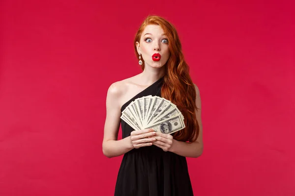 Luxury, beauty and money concept. Amazed silly good-looking young redhead woman in black stylish dress folding lips and looking surprised as winning cash, receive prize, stand red background