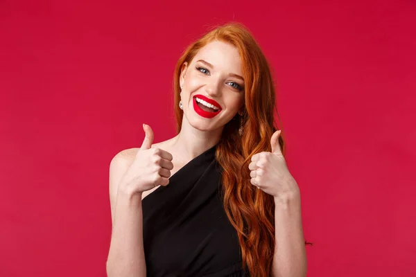 Közelkép portré boldog és elégedett vörös hajú európai nő ajánlom jó hely, viseljen fekete ruhát, party smink, mutasd hüvelykujj-up jóváhagyásban, mosolygós, mint a teljesítmény, piros háttér — Stock Fotó