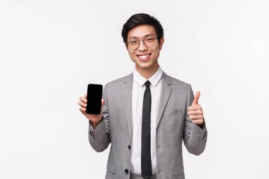Gri takım elbiseli yakışıklı Asyalı adamın belden yukarı portresi, cep telefonu ekranını ve baş parmaklarını göster, yeni uygulamayı onayla, satın alma önerisi, beyaz arka plan