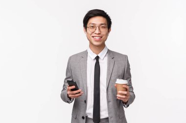 Ofis çalışanları, iş ve yaşam tarzı konsepti. Başarılı Asyalı takım elbiseli genç iş adamının belden yukarı portresi, kablosuz kulaklıklar, akıllı telefon ve paket kahve, gülümseyen kamera.