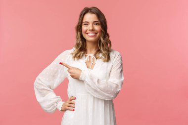 Pleasant good-looking smiling blond woman with blond short wavy hair, white tender dress, looking camera and pointing finger left to show you best price, good offer, promo concept, pink background clipart