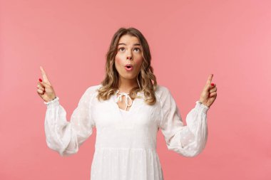 Heyecanlı, beyaz elbiseli sarışın, heyecanlı dudakları kıvrılmış nefes kesici, zevkli manzaralı, pembe arka planlı, nefes kesici bir şeyi işaret eden güzel beyaz bir kadın.