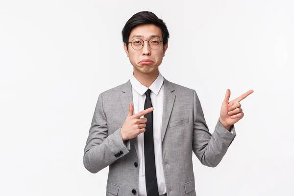 Retrato en la cintura del joven empresario asiático guapo impresionado, gerente de oficina en traje gris y corbata, señalando los dedos a la derecha, asiente con la cabeza en aprobación o acuerdo, como idea, fondo blanco —  Fotos de Stock