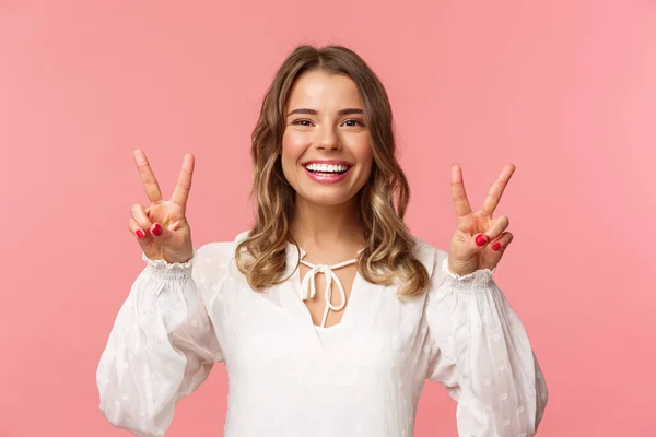 Primer plano retrato de optimista kawaii joven rubia con sonrisa radiante encantadora, mostrando signos de paz y mirando a la cámara con actitud positiva, disfrutando de la primavera, vestido blanco, fondo rosa — Foto de Stock