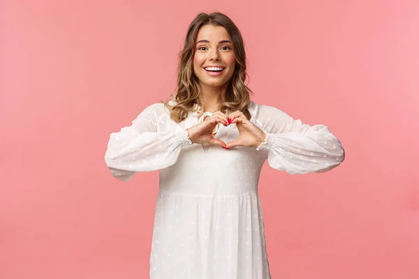 Conceito de beleza, moda e mulheres. Romântico apaixonado loiro europeu fêmea em vestido bonito branco, fazer sinal de coração perto do peito e sorrindo para a câmera animado, mostrar simpatia — Fotografia de Stock