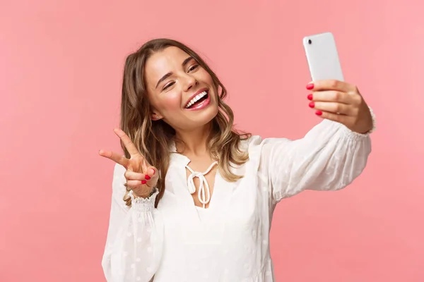 Närbild porträtt av glada vackra, feminina blond flicka i vit klänning, ta selfie på mobiltelefon, göra kawaii fred tecken medan ta foto, fånga våren ögonblick, rosa bakgrund — Stockfoto