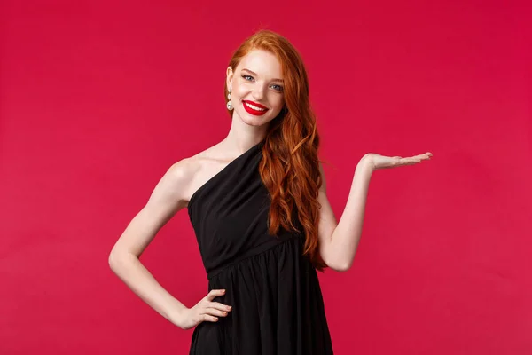 Retrato de mujer pelirroja joven elegante en lujoso vestido negro que presenta algo mantenga el producto en el brazo sobre el fondo rojo, introducir el servicio de la empresa, cámara sonrisa, mostrar promo — Foto de Stock