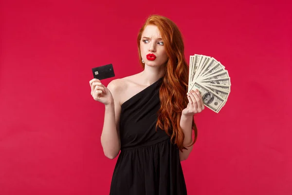 Concepto de lujo, belleza y dinero. Retrato de la joven pelirroja indecisa e insegura pensando qué hacer con un montón de dólares en efectivo en sus manos, mirando a la tarjeta de crédito vacilante — Foto de Stock