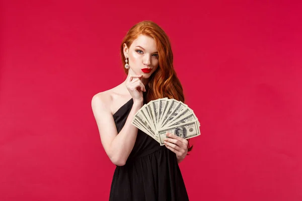 Luxury, beauty and money concept. Portrait of dubious and scheming good-looking young redhead woman in stylish black dress have interesting idea what buy on this cash dollars