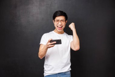 Teknoloji, aletler ve insan konsepti. Mmorpg online mobil oyununda zafer kutlayan mutlu Asyalı adamın portresi, neşeli bir yumruk, akıllı telefon atari şampiyonu.