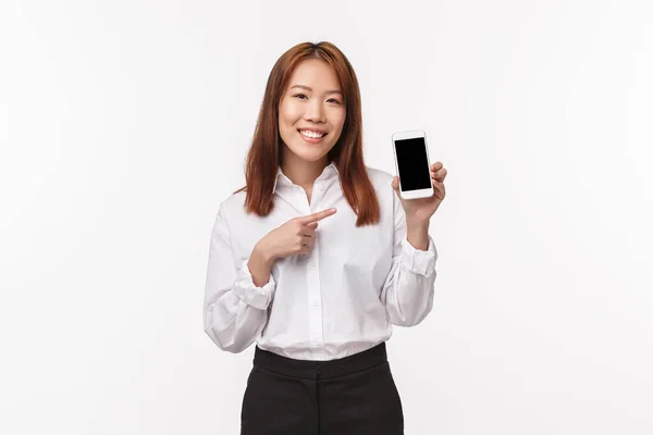 Porträt einer eleganten asiatischen Dame in weißem Hemd, die Ratschläge gibt, wo man Produkt, welche App herunterladen kann, Handy hält, Smartphone zeigt und Kamera lächelt, Studiohintergrund — Stockfoto