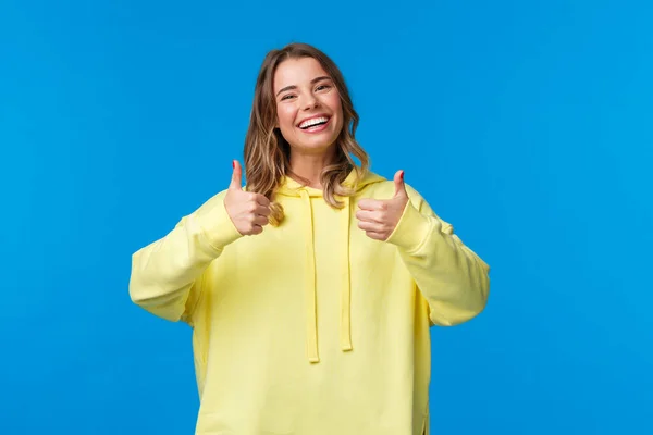 Sounds good Im in. Cheerful and optimistic good-looking young female blonde in yellow hoodie, show thumbs-up in approval, smiling nod acceptingly, like idea, blue background — 스톡 사진
