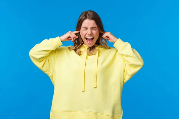 Mujer rubia molesto y perturbado molesto cerrar los oídos y gritar como escuchar ruidoso ruido molesto y perturbador, sentir la incomodidad de la sala de estar, de pie fondo azul —  Fotos de Stock