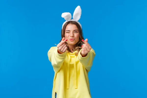 Feiertage, Traditionen und Festkonzept. Dummes und nettes, glückliches blondes Mädchen, das Spaß hat, koreanische Herzgeste mit Fingern zeigt und Küsse schickt, in Hasenohren steht, gelben Kapuzenpullover — Stockfoto