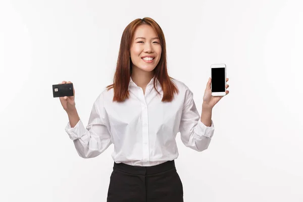 Porträt einer fröhlichen jungen Asiatin mit stolzem, strahlenden Lächeln, die Smartphone und Kreditkarte zeigt, cooles Frühlingsoutfit oder Sommerurlaub im Internet-Shop mit Geld auf Rechnung bestellt — Stockfoto