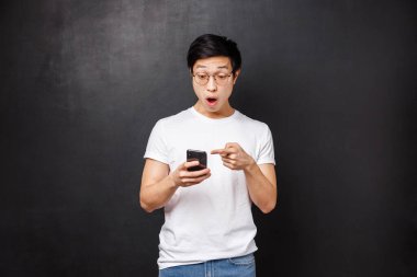 Teknoloji, aletler ve insan konsepti. Etkilenmiş ve afallamış genç Asyalı adam online mağazada harika bir ürün gördü, cep telefonu ekranını işaret etti, şaşırmış bir yüz, siyah arka plan