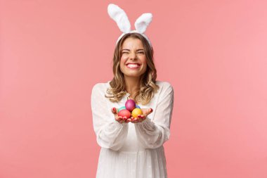 Tatiller, bahar ve parti konsepti. Güzel gülüşü olan, elinde boyalı yumurta tutan, Paskalya kıyafeti giyen, tavşan kulaklı, beyaz elbiseli, güzel bir sarışın kadının portresi.