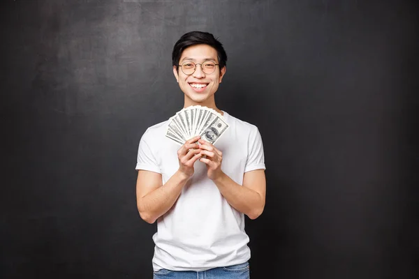 Retrato de afortunado satisfecho joven rico asiático estudiante masculino sosteniendo un montón de dinero en efectivo, fan de los dólares sonriendo feliz y contento como premio ganador, recibir recompensa o su primer cheque de pago, fondo negro — Foto de Stock