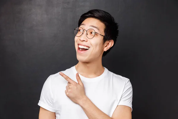 Närbild porträtt av entusiastisk leende asiatisk man i vit t-shirt och glasögon står mot svart bakgrund, tittar och pekar övre vänstra hörnet på cool awesome produkt — Stockfoto