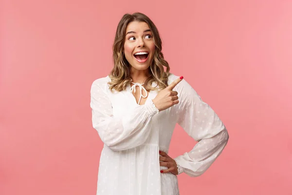 Entusiástico, mulher loira atraente feliz em vestido bonito branco, boca aberta animado e alegre olhando, apontando canto superior direito como melhores descontos de preços sempre, stand pink background — Fotografia de Stock