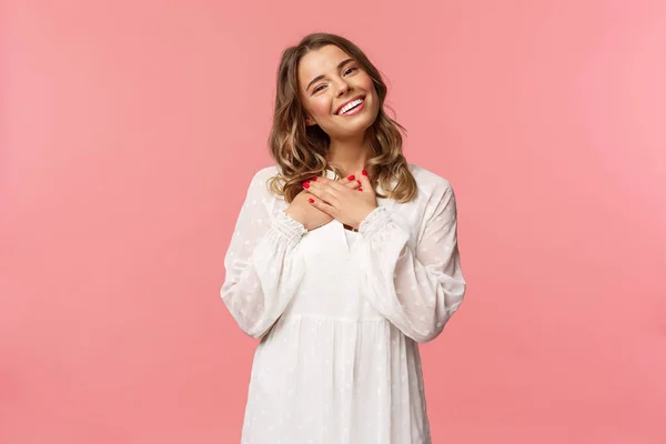 Belleza, moda y concepto de mujer. Retrato de mujer joven rubia femenina en vestido blanco lindo, mantenga los brazos presionados al corazón y suspirando de admiración y deleite, siéntase agradecida, fondo rosa — Foto de Stock