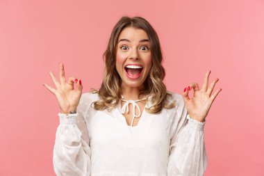 Heyecanlı, neşeli, beyaz elbiseli, neşeli sarışın kızın portresi mutlu bir gülümsemeyle iyi işaretler gösteriyor, müzik festivali için heyecanlı, harika bir film derecesi var, ürün öneriyor.