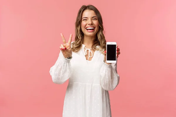 Porträtt av kawaii optimistisk och glad ung flicka i vit klänning, visa mobiltelefon display och fred tecken, skrattar känsla glad och glad att dela fantastisk app, användbar länk, rosa bakgrund — Stockfoto