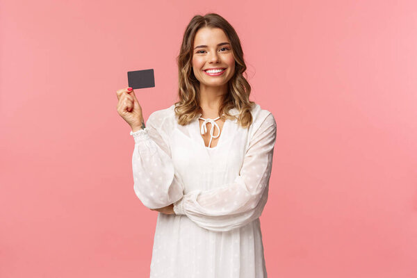 Портрет симпатичной белокурой блондинки в белом платье, показать кредитную карту с удовлетворённым выражением лица, улыбающаяся камера, рекомендовать банковские услуги, использовать оплату онлайн, розовый фон
