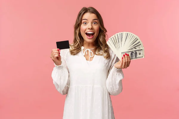 Portret podekscytowanej szczęśliwej blondynki w białej sukience, wygrywającej pieniądze, postawionej na dobry zakład, zawartej transakcji, trzymającej dolary i kartę kredytową, uśmiechniętej do kamery, różowe tło — Zdjęcie stockowe