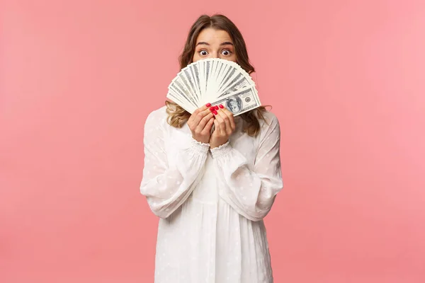 Retrato de surpreendido e animado bonito menina loira feminina em vestido branco, segurando dólares sobre o rosto, olhando de baixo do dinheiro para a câmera com expressão surpresa, stand pink background — Fotografia de Stock