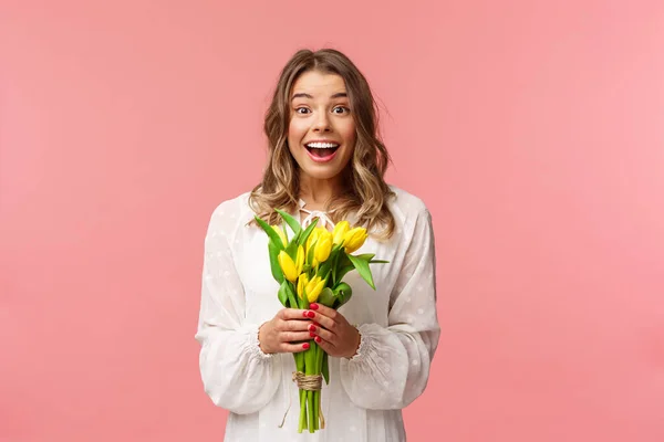 Vacaciones, belleza y concepto de primavera. Retrato de excitada y sorprendida joven hermosa rubia en vestido blanco, la celebración de tulipanes amarillos, recibir flores como regalo, oliendo divertido, fondo rosa — Foto de Stock