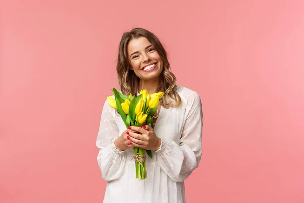 Ünnepek, szépség és tavasz. Portré szép kaukázusi szőke lány fehér ruhában, mosolygós vidám, gazdaság sárga tulipán, amelynek tökéletes romantikus dátum virágokkal ajándékba, rózsaszín háttér — Stock Fotó
