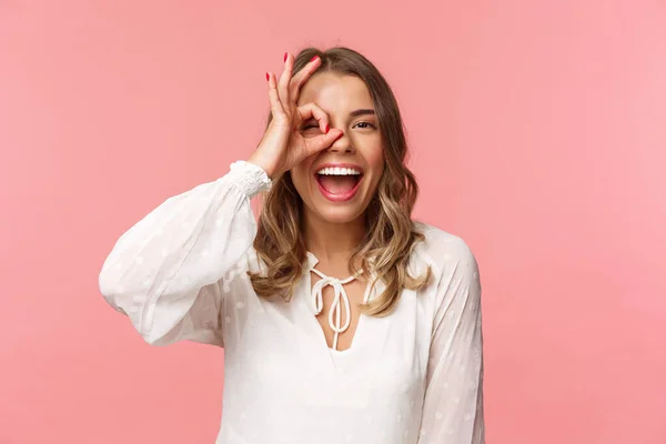 Retrato de cerca de la perfecta chica rubia guapa en vestido blanco, mirando a través de signo de bien y sonriendo, garantía de excelente calidad, aprobar o como algo, de pie fondo rosa —  Fotos de Stock