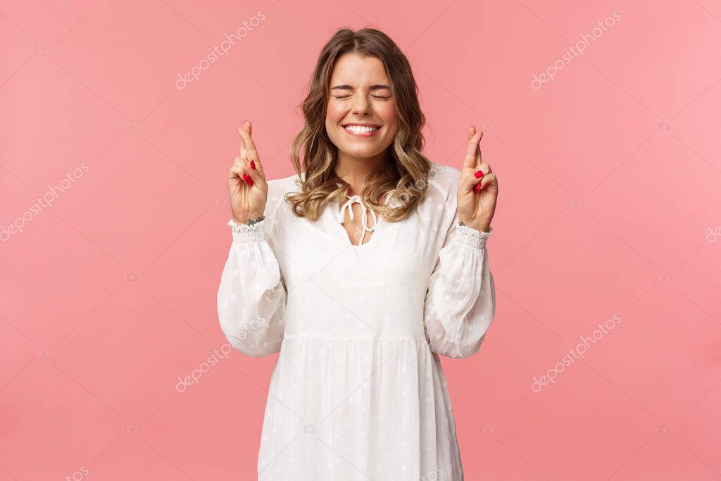 Portrait of hopeful lucky cute blond girl in white dress praying, anticiapte miracle, making wish to win, cross fingers good luck, smiling and close eyes and pleading god, pink background