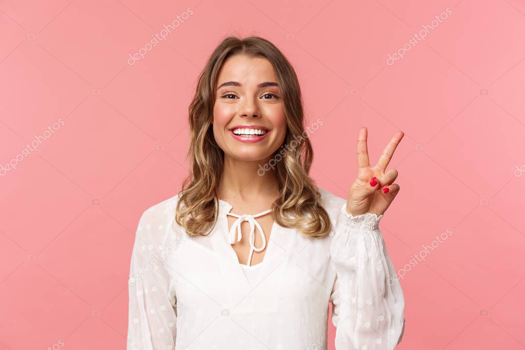 Close-up portrait of romantic lovely smiling girl with blond short hair, wearing white dress, show peace sign enjoying spring, grinning and look camera over pink background