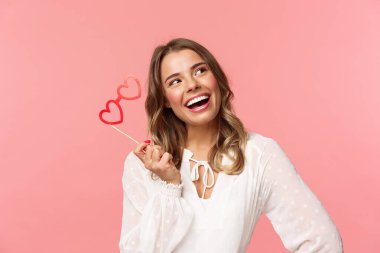 Bahar, mutluluk ve kutlama konsepti. Güzel sarışın kızın yakın plan portresi, kalp şeklinde bir maske takıyor ve hayal kuruyor, mutlu bir gülümsemeyle düşünceli bir şekilde yukarı bakıyor.