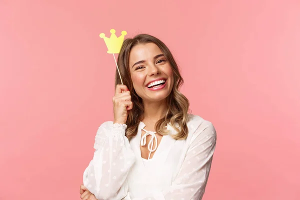 Concepto de primavera, felicidad y celebración. Retrato de cerca de la encantadora chica rubia sonriente y encantadora sosteniendo la pequeña corona de la reina en un palo, riendo alegremente, sintiéndose empoderada y feliz, fondo rosa — Foto de Stock