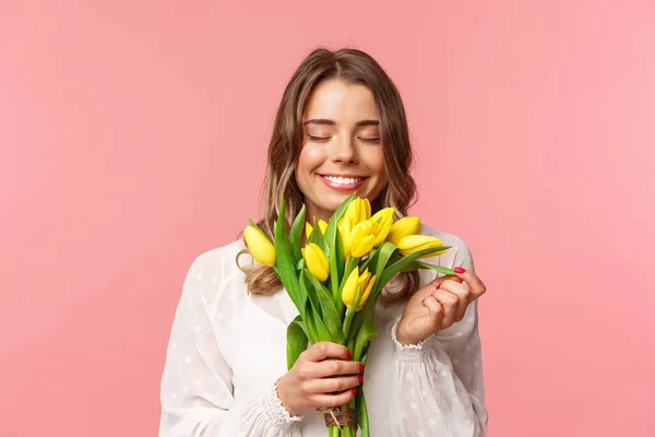 Concepto de primavera, felicidad y celebración. Retrato de cerca de una chica rubia encantadora y romántica oliendo a hermosos tulipanes amarillos, ojos cerrados y sonriente, fondo rosa de pie — Foto de Stock