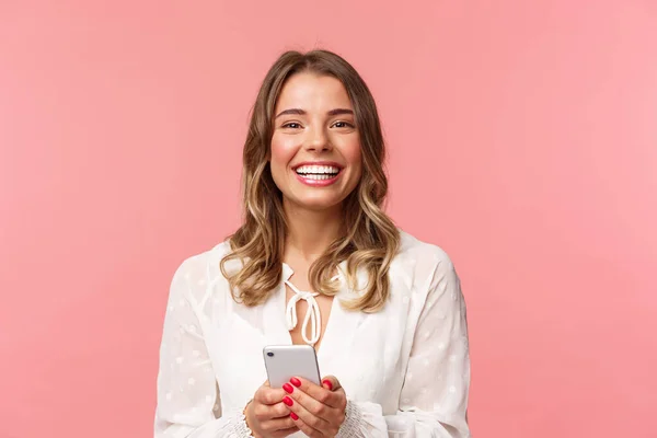 Retrato de cerca de la encantadora y encantadora chica rubia usando el teléfono móvil, enviando mensajes de texto a un amigo, ordene en la tienda en línea usando la aplicación para teléfonos inteligentes, riendo y sonriendo complacido, de pie fondo rosa —  Fotos de Stock