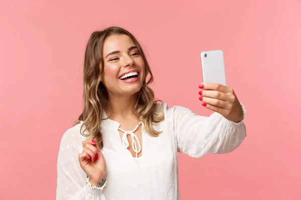 Крупный план портрет веселой оптимистичной улыбающейся блондинки, одетой в белое платье, смеющейся как видеозапись, звонящей подруге по мобильному приложению, делающей фото, селфи на смартфоне, розовый фон — стоковое фото