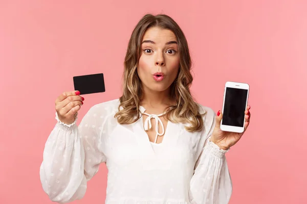 Concepto de finanzas, compras y tecnología. Retrato de cerca de la chica linda rubia excitada en vestido blanco, labios plegables mirada divertida cámara como muestra la tarjeta de crédito y el teléfono móvil, fondo rosa — Foto de Stock