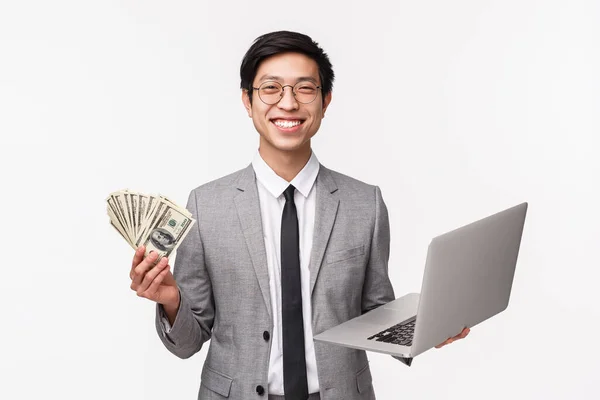 Cintura de feliz, contento sonriente joven asiático en traje gris sosteniendo dólares, hacerse rico, el uso de la computadora portátil como explicar cómo invertir en la empresa financiera, ganar premio en línea, fondo blanco — Foto de Stock
