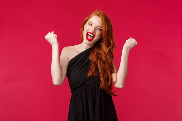 Портрет успешной молодой довольной рыжей девушки, побеждающей, чувствующей себя накаченной, поднимающей кулаки, как чемпионка, говорящей да поющей и танцующей победу, торжествующей в черном платье на красном фоне — стоковое фото