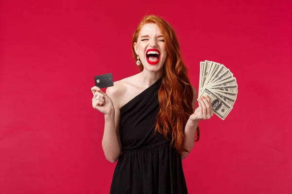 Concepto de lujo, belleza y dinero. Retrato de triunfar mujer pelirroja atractiva afortunada en vestido elegante negro gritando de placer y alegría, ganar premios en efectivo, mantener la tarjeta de crédito y dólares — Foto de Stock