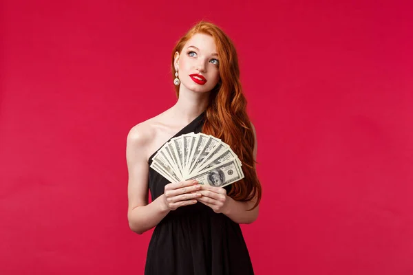 Luxury, beauty and money concept. Dreamy good-looking redhead woman with lots cash, become rich after receiving prize or reward, look up dreaming, thinking how waste income, red background
